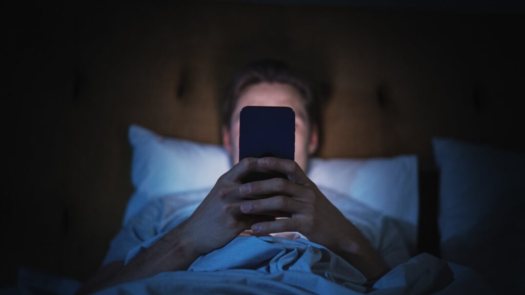 Osoba leżąca w łóżku trzyma smartfon i patrzy w ekran w ciemnym pomieszczeniu.